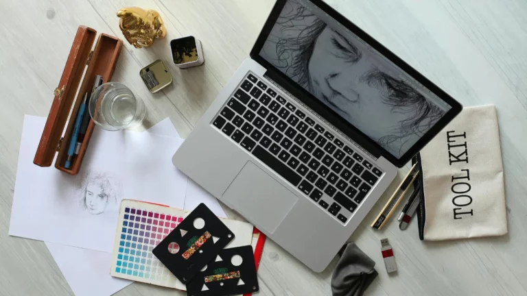 A laptop with a digital portrait on-screen, surrounded by sketching tools, a colour palette, and drawing paper.