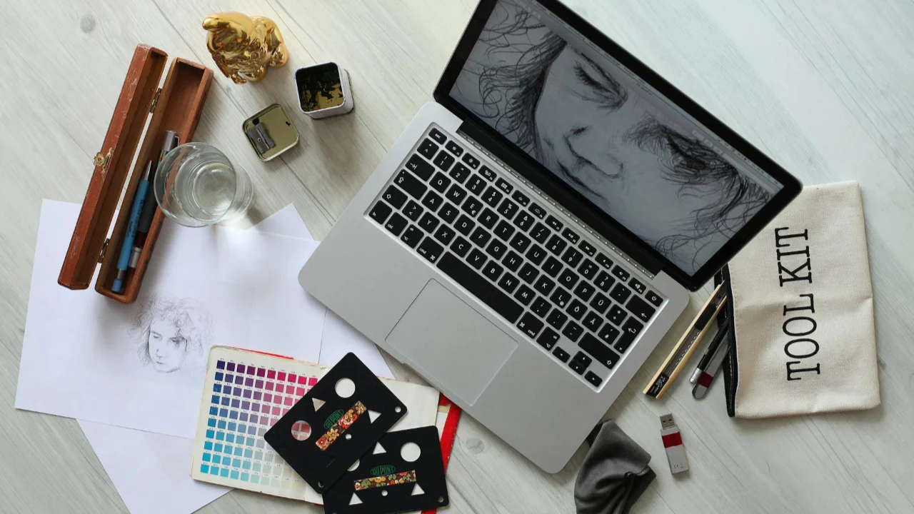 A laptop with a digital portrait on-screen, surrounded by sketching tools, a colour palette, and drawing paper.